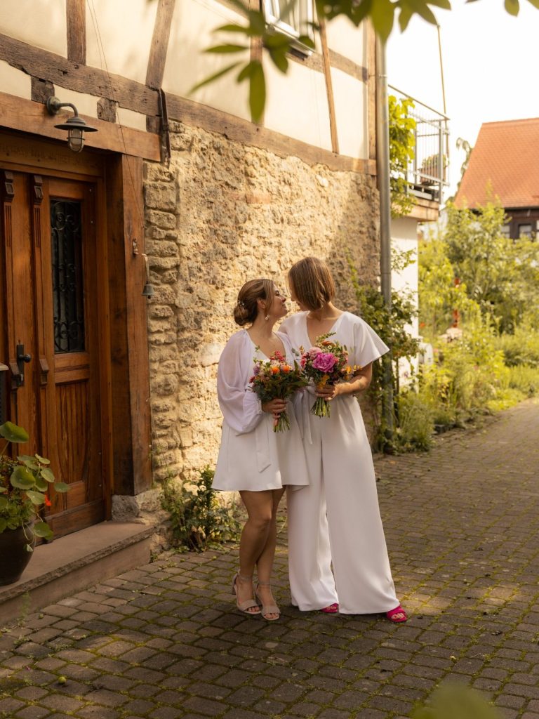 Hochzeit Zwei Bräute stehen auf einem gepflasterten Weg vor einem Fachwerkhaus und halten Blumen.
