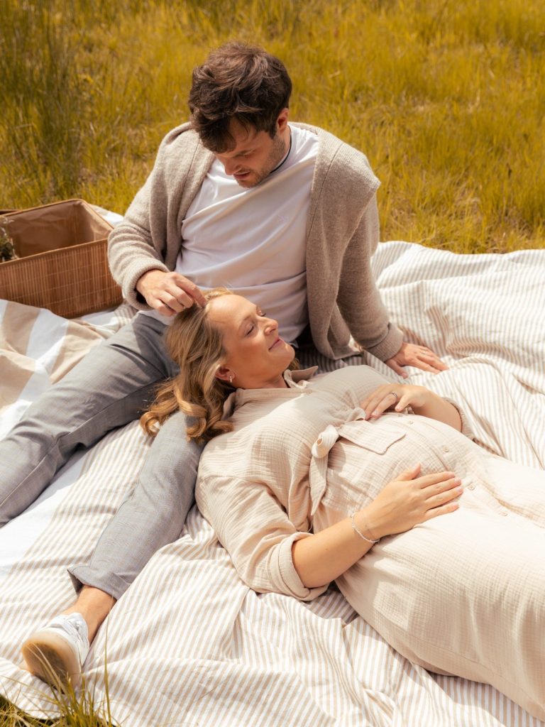 Paar entspannt auf einer Decke in der Natur, schwangere Frau liegt, er spielt mit ihrem Haar.